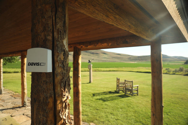 White Air Quality Monitor by Davis Instruments attached to a wooden porch post in a green outdoor setting.
