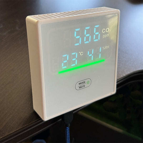 White indoor air quality monitor by Sper Scientific displaying CO2, humidity, and temperature readings, with a green indicator bar.