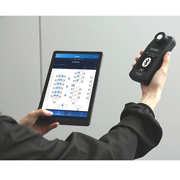 Light meter by Sekonic paired with a tablet showing a layout, held in a person's hands.
