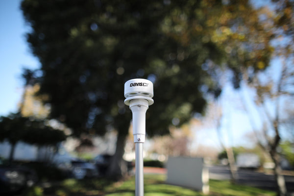 Weather monitoring sensor by Davis Instruments with a white cylindrical body and silver pole, set against a blurred outdoor background.
