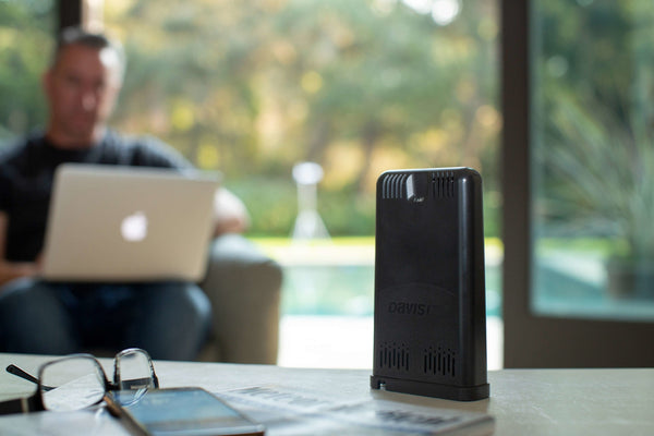 Black weather data hub by Davis Instruments with vent details, shown on a table with a laptop in the background.
