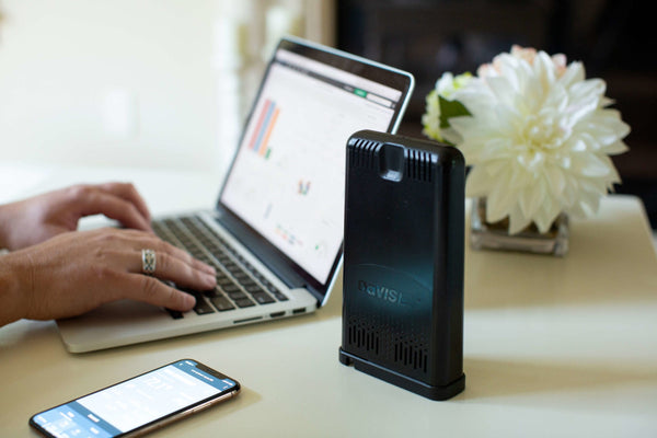 Black WeatherLink Live data collection device by Davis Instruments displayed on a table with a laptop, phone, and vase of flowers.