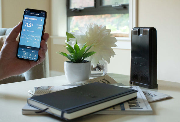 Black WeatherLink Live data collection device by Davis Instruments on a table next to a phone displaying a weather app.
