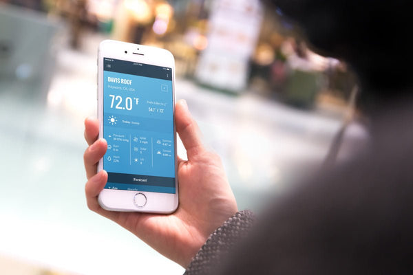 Weather app interface by Davis Instruments showing a sunny forecast on a smartphone held in hand.