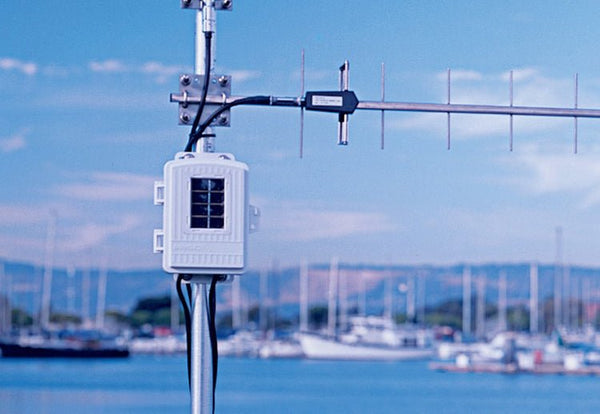 White wireless long-range repeater with solar power by Davis Instruments mounted on a silver pole against a blurred water and boat background.
