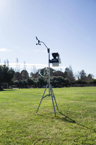Weather station by Davis Instruments with anemometer, thermometer, rain collector on metal tripod in grassy field.
