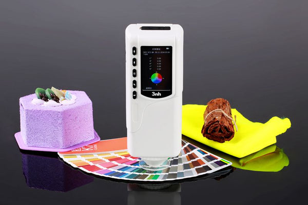 White precision colorimeter by 3nh standing on a colorful chart, with a yellow cloth and cake nearby.