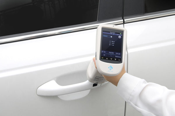 White spectrophotometer by 3nh, displaying color measurement metrics, being held against a white car door.